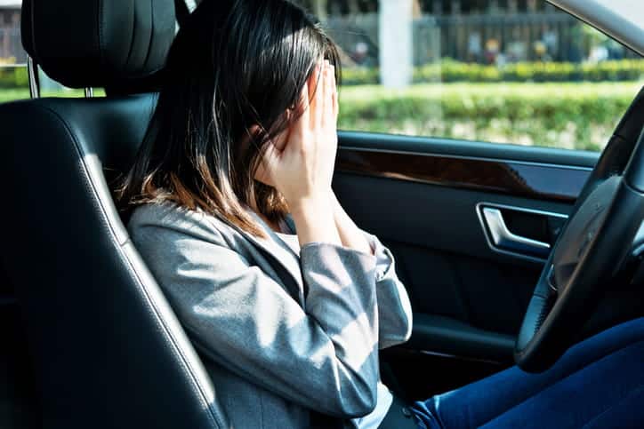 A Woman Sitting In The Driver'S Seat Of A Car, Covering Her Face With Both Hands In A Gesture Of Distress Or Overwhelm.