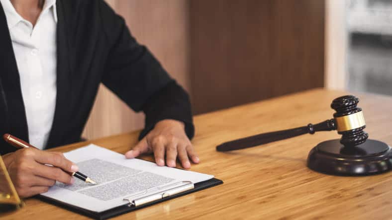 A Legal Professional In A Black Suit Sitting At A Wooden Desk, Signing Or Reviewing Documents On A Clipboard With A Judge'S Gavel In The Background.