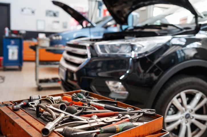 A Close-Up Of A Tray Filled With Various Mechanic Tools In The Foreground, With A Black Car'S Open Hood And An Auto Repair Shop Interior Blurred In The Background