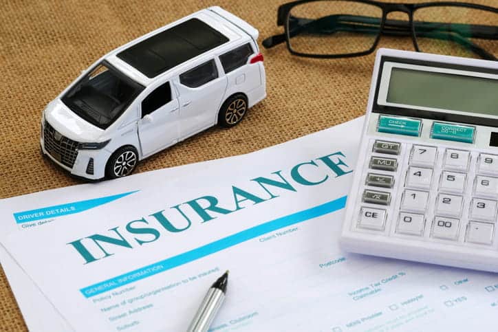 An Overhead View Of A White Car Insurance Form On A Desk, Accompanied By A White Toy Car, A Calculator, A Pen, And A Pair Of Eyeglasses.