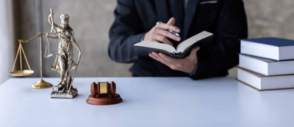 A Legal Professional In A Suit Reviewing A Book At A Desk Featuring A Lady Justice Statue, A Wooden Judge'S Gavel, And A Stack Of Law Books.