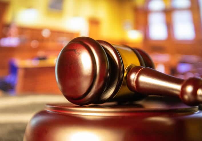 A Close-Up, Low-Angle Shot Of A Wooden Judge'S Gavel Resting On Its Sound Block, With A Blurred Law Office Or Courtroom Interior In The Background.