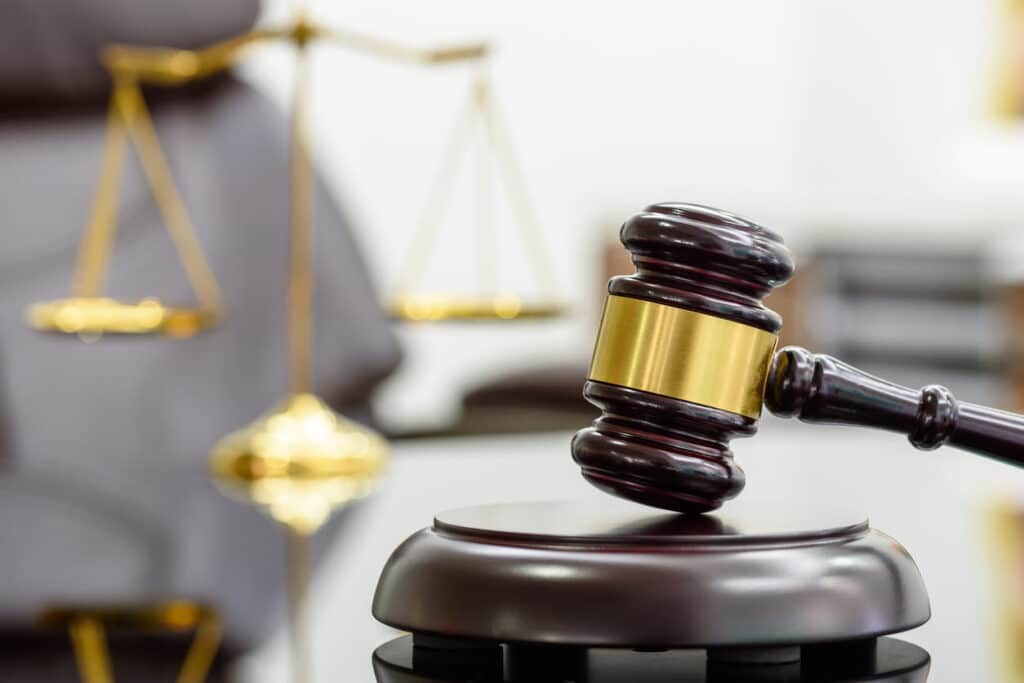 A Close-Up Of A Dark Wooden Judge'S Gavel With Gold Accents On A Sound Block, With The Golden Scales Of Justice Blurred In The Background.