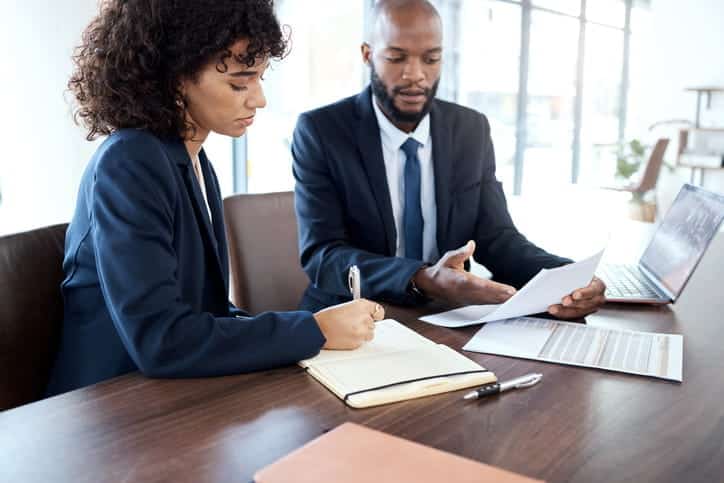 A Male And Female Attorney In Professional Suits Sitting At A Conference Table, Reviewing Paperwork And Taking Notes During A Legal Consultation.