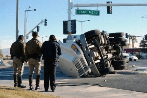 A Large White Semi-Truck Lies On Its Side After Rolling Over At A City Intersection Under A &Quot;Lake Mead&Quot; Street Sign. Three Law Enforcement Officers In Uniform Stand On The Sidewalk In The Foreground, Surveying The Scene. The Truck'S Undercarriage And Wheels Are Exposed.