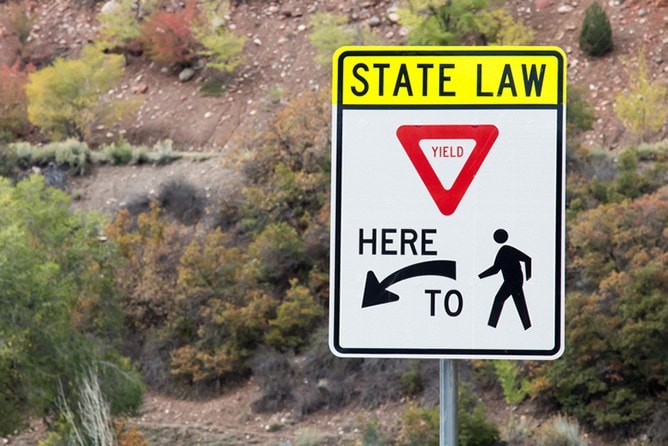 Leyes De Derecho De Paso Y Falta De Ceder El Paso En Nevada 2 A Vertical Rectangular Road Sign With A Yellow Header That Reads &Quot;State Law.&Quot; Below, It Features A Red And White Yield Triangle And The Text &Quot;Here To&Quot; With An Arrow Pointing Left Toward A Black Silhouette Of A Person Walking.