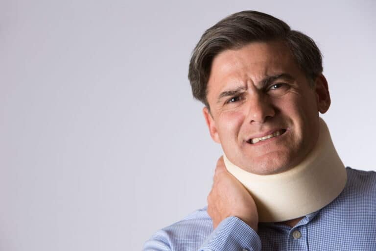 Fingir Lesiones En Accidentes De Auto 1 Retrato De Un Hombre Con Expresión De Dolor Usando Un Collarín Ortopédico De Espuma Mientras Sostiene Su Cuello Con La Mano Sobre Un Fondo Gris Claro.