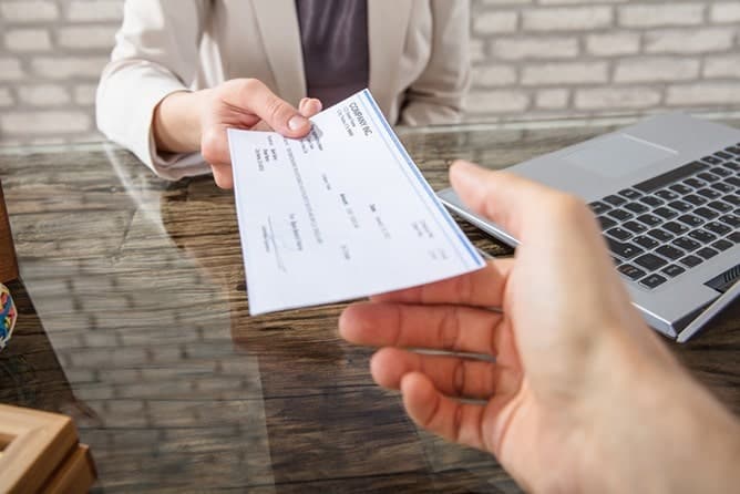 Una Persona Con Traje Formal Entrega Un Cheque De Pago Único Sobre Un Escritorio De Cristal A Otra Persona, Con Una Computadora Portátil A Un Costado.