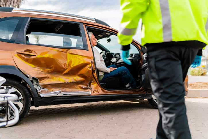 Un Paramédico Con Chaqueta De Seguridad Fluorescente Se Acerca A Un Vehículo Naranja Con Daños Severos Por Colisión Lateral, Donde Un Conductor Mayor Permanece Sentado Tras El Accidente.
