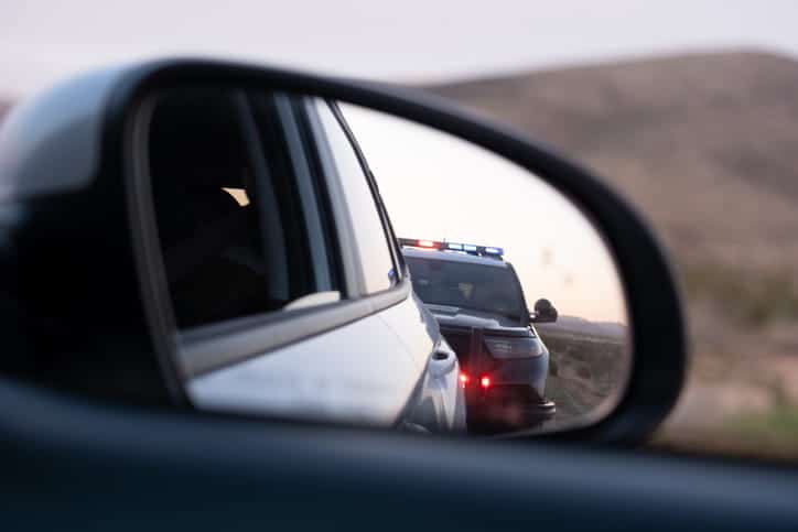 ¿Puede La Policía Detenerte Si No Has Hecho Nada Malo? 1 Vista A Través Del Espejo Lateral De Un Coche Que Muestra Una Patrulla De Policía Con Luces Rojas Y Azules Encendidas, Deteniéndose Detrás En Una Carretera Al Atardecer.