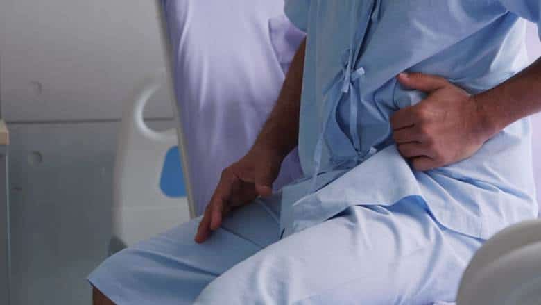 Signos De Lesión Interna Después De Un Accidente De Auto 1 Paciente Con Bata Azul De Hospital Sentado En Una Cama Mientras Se Sujeta El Costado Izquierdo Del Abdomen Con Gesto De Dolor.