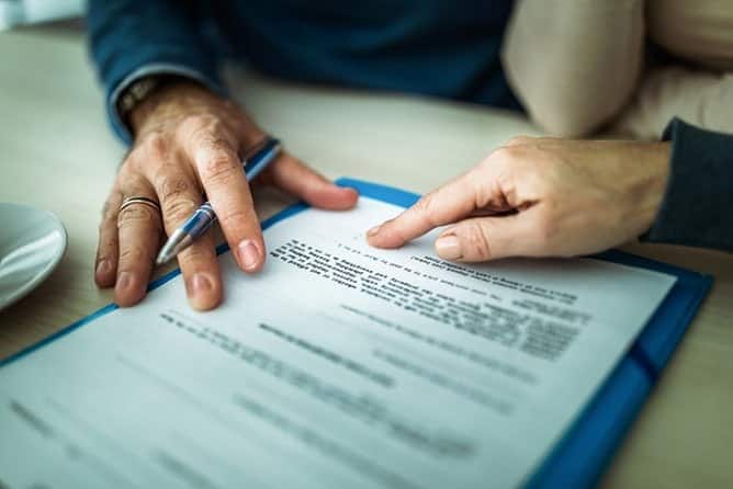 Cómo Tratar Con Los Ajustadores De Seguros De Auto Y Mantenerte Un Paso Adelante 1 Primer Plano De Dos Personas Revisando Un Contrato De Acuerdo Sobre Una Carpeta Azul, Con Una Mano Sosteniendo Un Bolígrafo Y El Dedo De Otra Persona Señalando Una Cláusula Específica Del Documento.