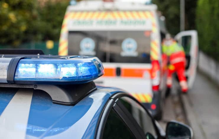 Lesiones Catastróficas En Paradise 1 Patrulla Policial Con Luces Azules Encendidas En Primer Plano Y Una Ambulancia Con Paramédico Atendiendo Una Emergencia Al Fondo, En El Lugar De Un Accidente Con Lesiones Catastróficas.