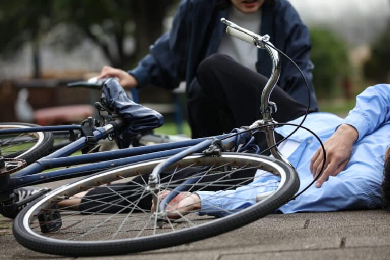 Abogado De Accidentes De Bicicleta En Henderson 1 Persona Tendida En El Suelo Tras Un Accidente De Bicicleta, Con Otra Persona Arrodillada A Su Lado En Un Entorno Exterior.