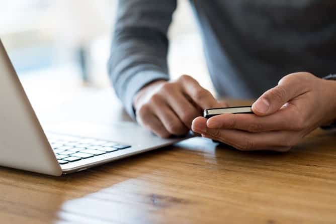Cómo Las Compañías De Seguros Investigan Tus Reclamos Por Accidente De Auto 1 Primer Plano De Las Manos De Una Persona Utilizando Un Teléfono Celular Sobre Un Escritorio De Madera, Al Lado De Una Computadora Portátil Abierta.